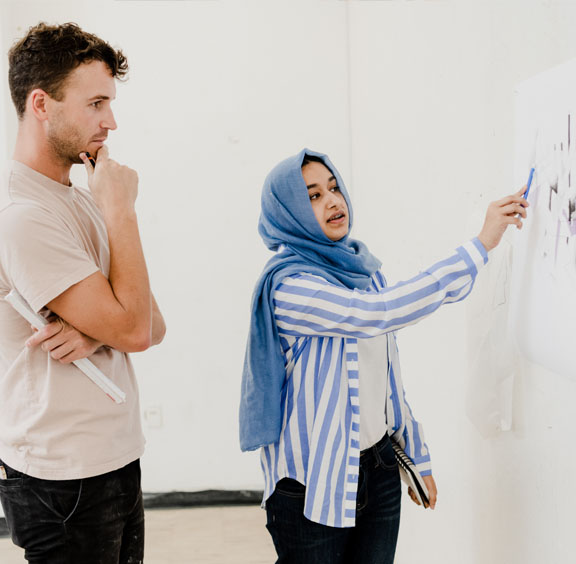 Example of a momentum moments photo. An architecture student and professor discuss designs drawn on a whiteboard. The student points at the board and is wearing a medium blue head scarf, a striped blue overshirt, a white shirt, and black pants. The professor stands with chin in hand and is wearing a light orange or beige shirt and black pants.